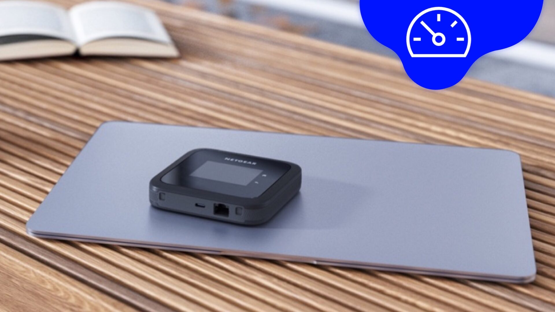Netgear Nighthawk M6 mobile Router liegt auf einem Macbook Air, das auf einem Holztisch steht. Am oberen rechten Rand des Bildes befindet sich eine Digital Republic Fluidform mit einem Speed-Icon
