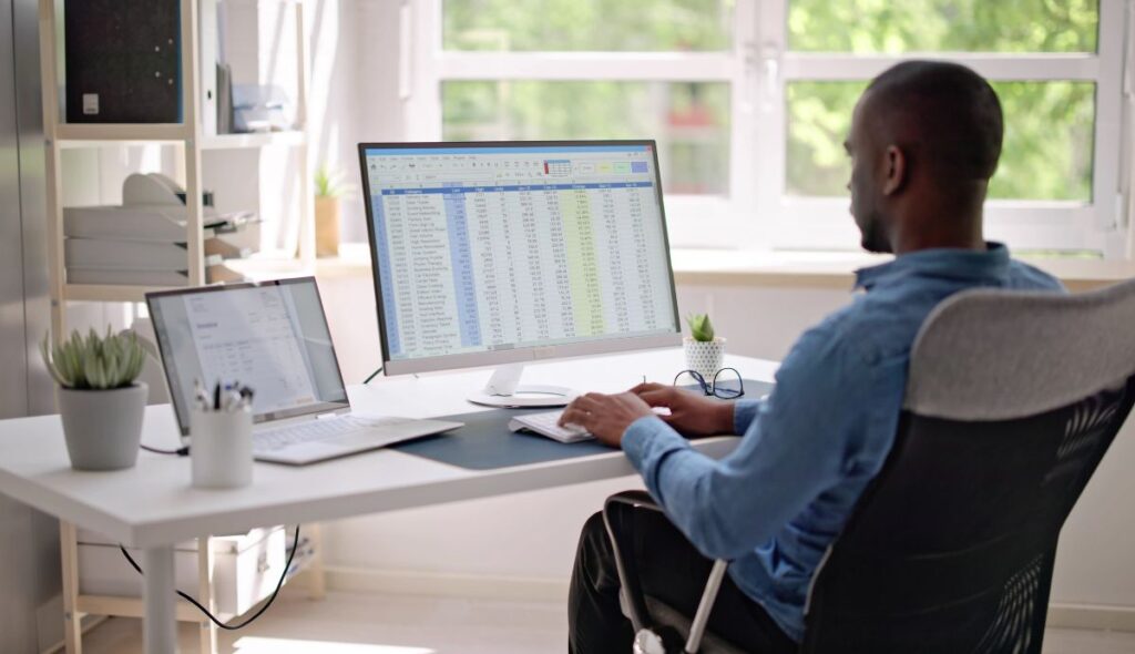 Mann sitzt mit aufgeklapptem Laptop an einem Schreibtisch und führt professionelle Arbeiten an einem externen Bildschirm mit einer externen Tastatur aus.